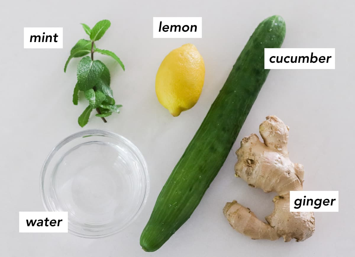 sprig of fresh mint, bowl of water, cucumber, ginger, and lemon on a white counter with text overlay describing ingredients.