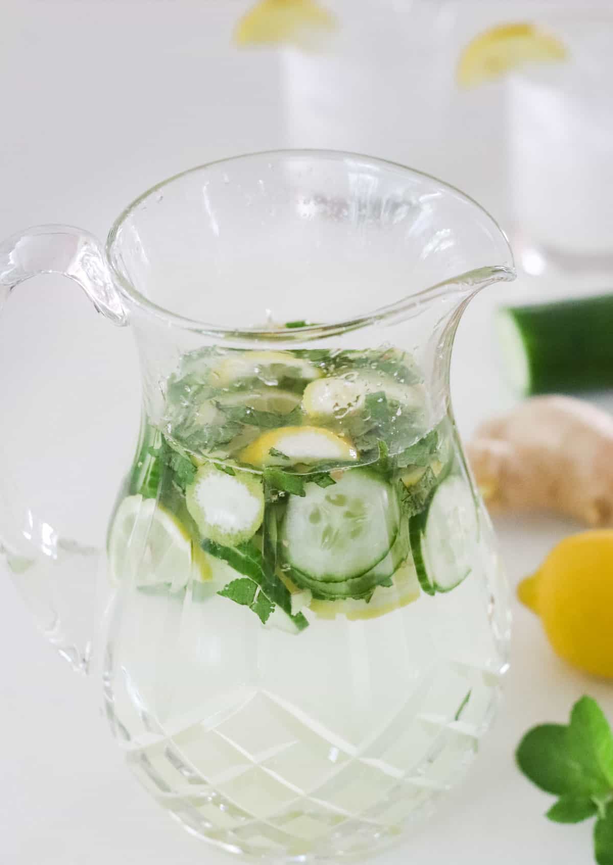 crystal pitcher with cucumber mint water next to lemon, mint, ginger, and cucumber.