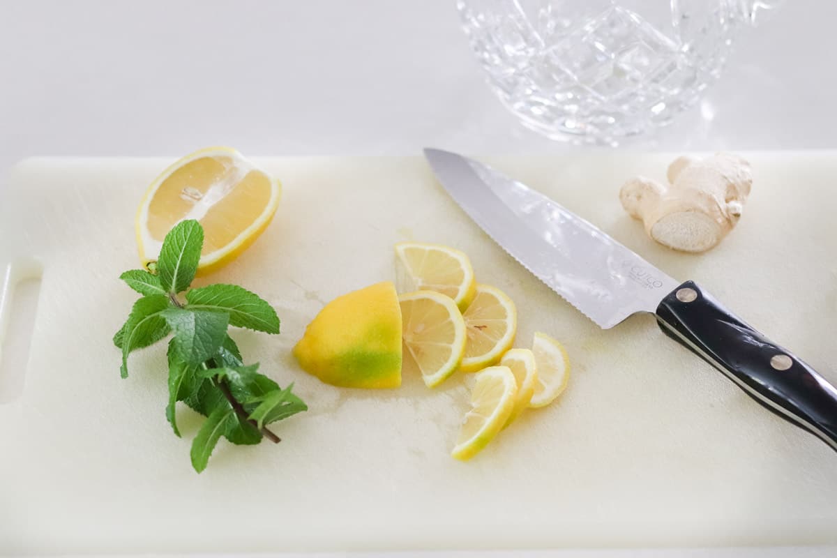 plastic cutting board with sprig of fresh mint, sliced lemon, ginger root, and a knife.