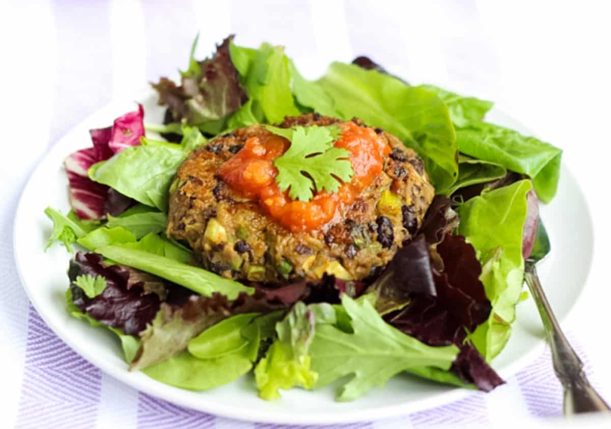 one black bean avocado burger topped with salsa and cilantro on a bed of mixed lettuce, silver fork, and white plate with purple and white striped towel underneath.