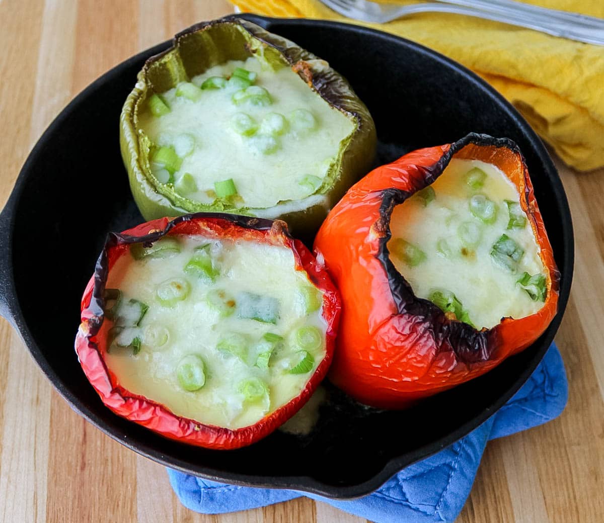 two red bell peppers and one green pepper filled with eggs, cheese, and green onions in a cast iron skillet on a blue hot pad on a wooden table.