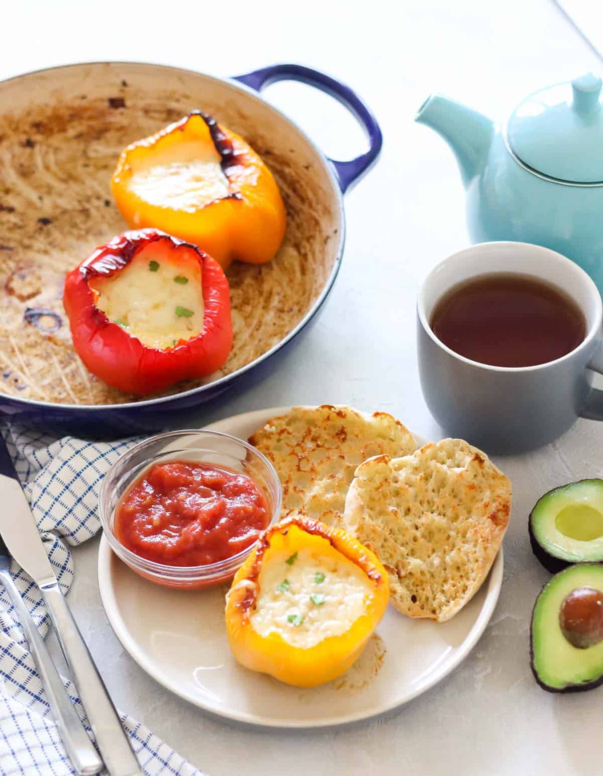 blue pot with breakfast stuffed peppers next to a teal tea pot, cup of tea, cut avocado, fork and knife, and a plate with yellow bell pepper stuffed with eggs and cheese, bowl of salsa, and English muffin.