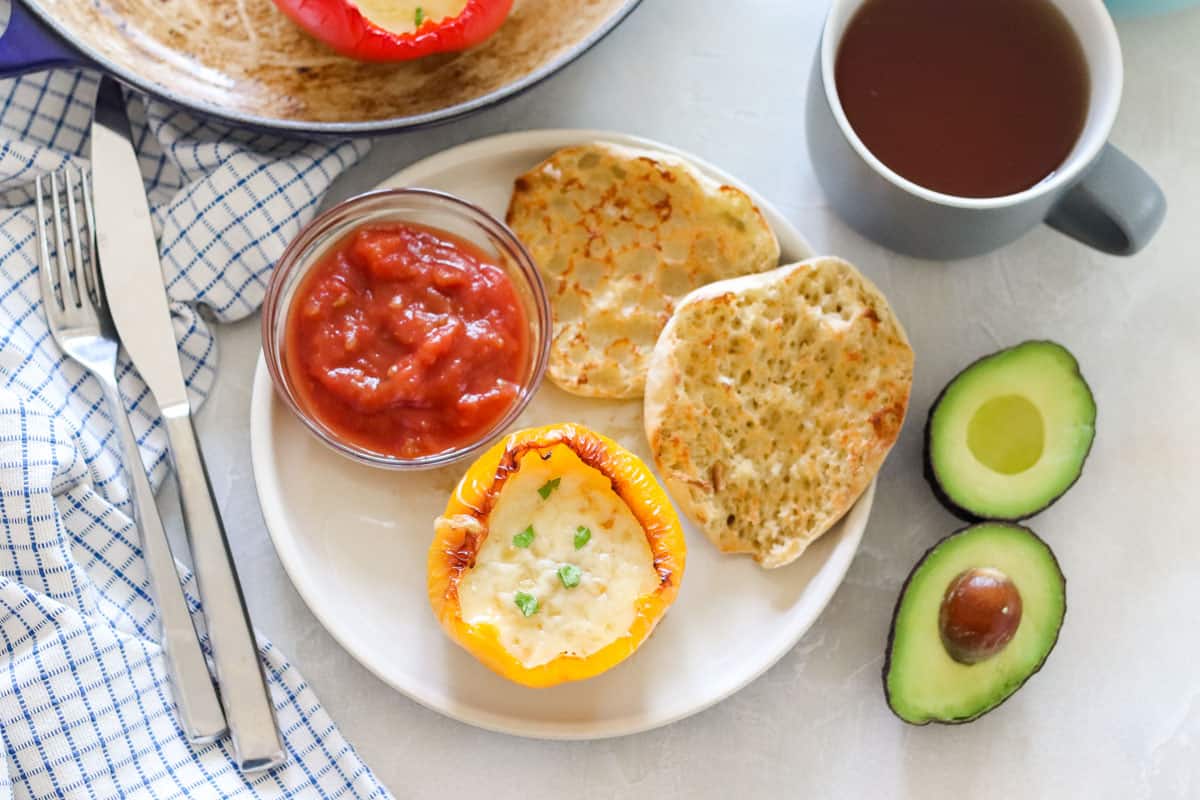 plate with breakfast stuffed bell pepper, bowl of salsa, and English muffin, next to napkin with silver fork and knife, cut avocado, and cup of tea.