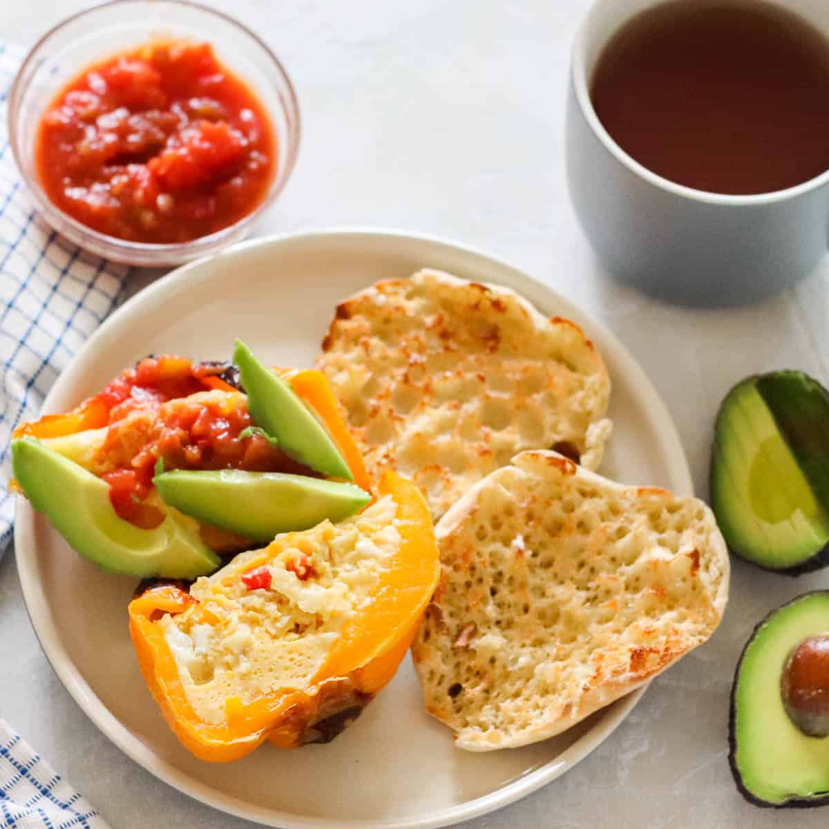 cut open egg stuffed bell pepper topped with salsa and avocado on a plate with an English muffin, next to a cut avocado, bowl of salsa, and cup of tea.