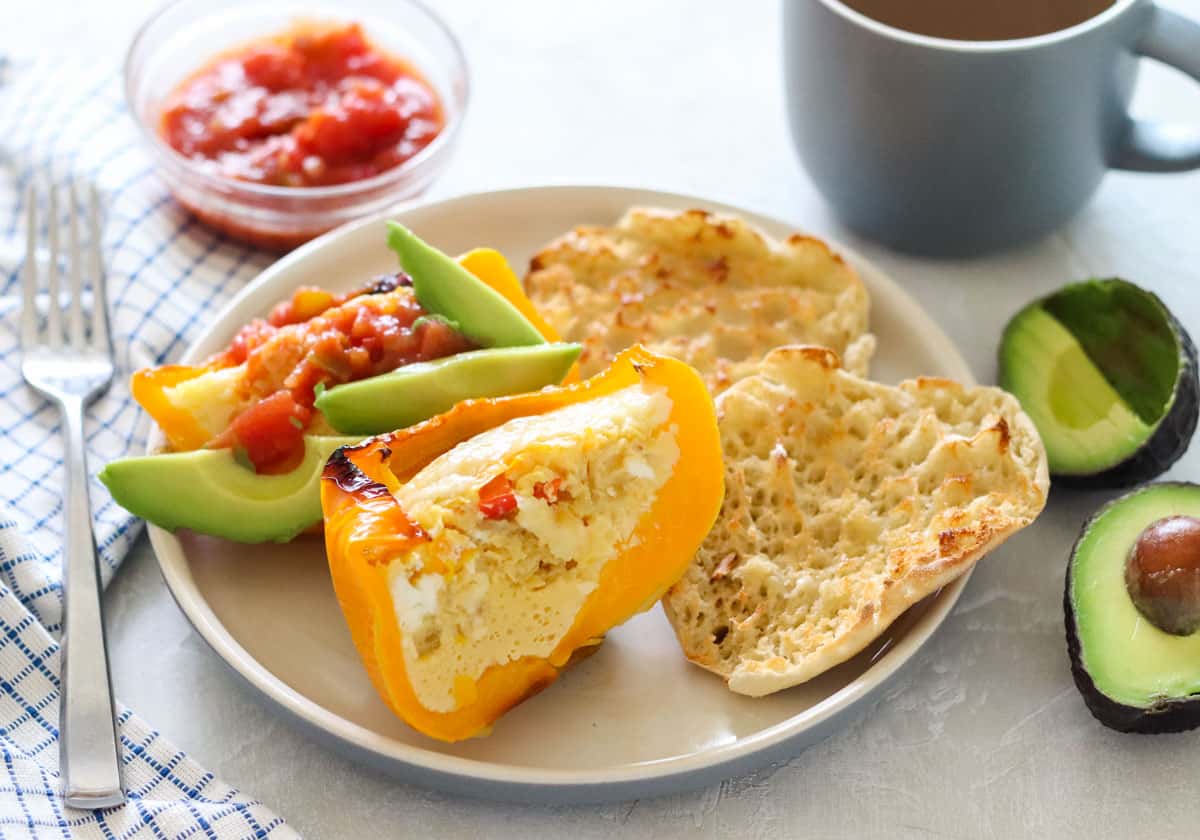 yellow bell pepper filled with eggs and cheese cut open on a plate, topped with avocado and salsa, next to english muffin and an avocado, fork, bowl of salsa, and tea.