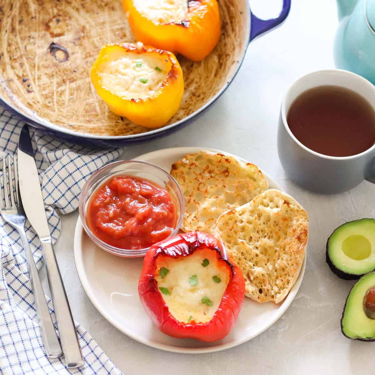 fork and knife, avocado, cup of tea, pot of stuffed breakfast peppers, and a red bell pepper stuffed with cheese and eggs on a plate next to english muffin and bowl of salsa.