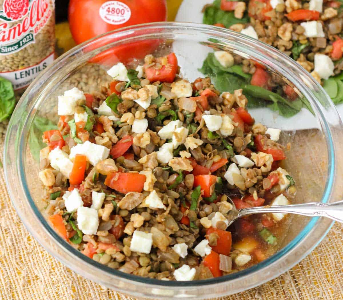 clear bowl with a silver spoon, cooked lentils, mozzarella, walnuts, tomatoes, and basil on a straw mat.