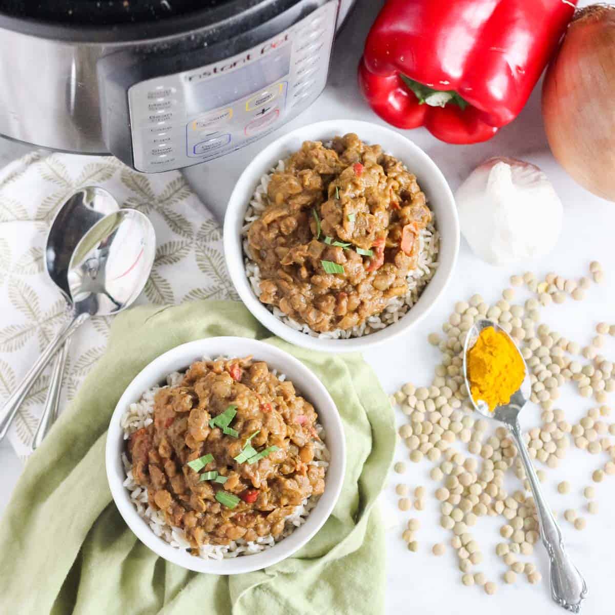 Instant Pot, two silver spoons, green napkins, white bowls with lentil curry and rice, spoon with turmeric, scattered green lentils, red bell pepper, garlic, and onion.