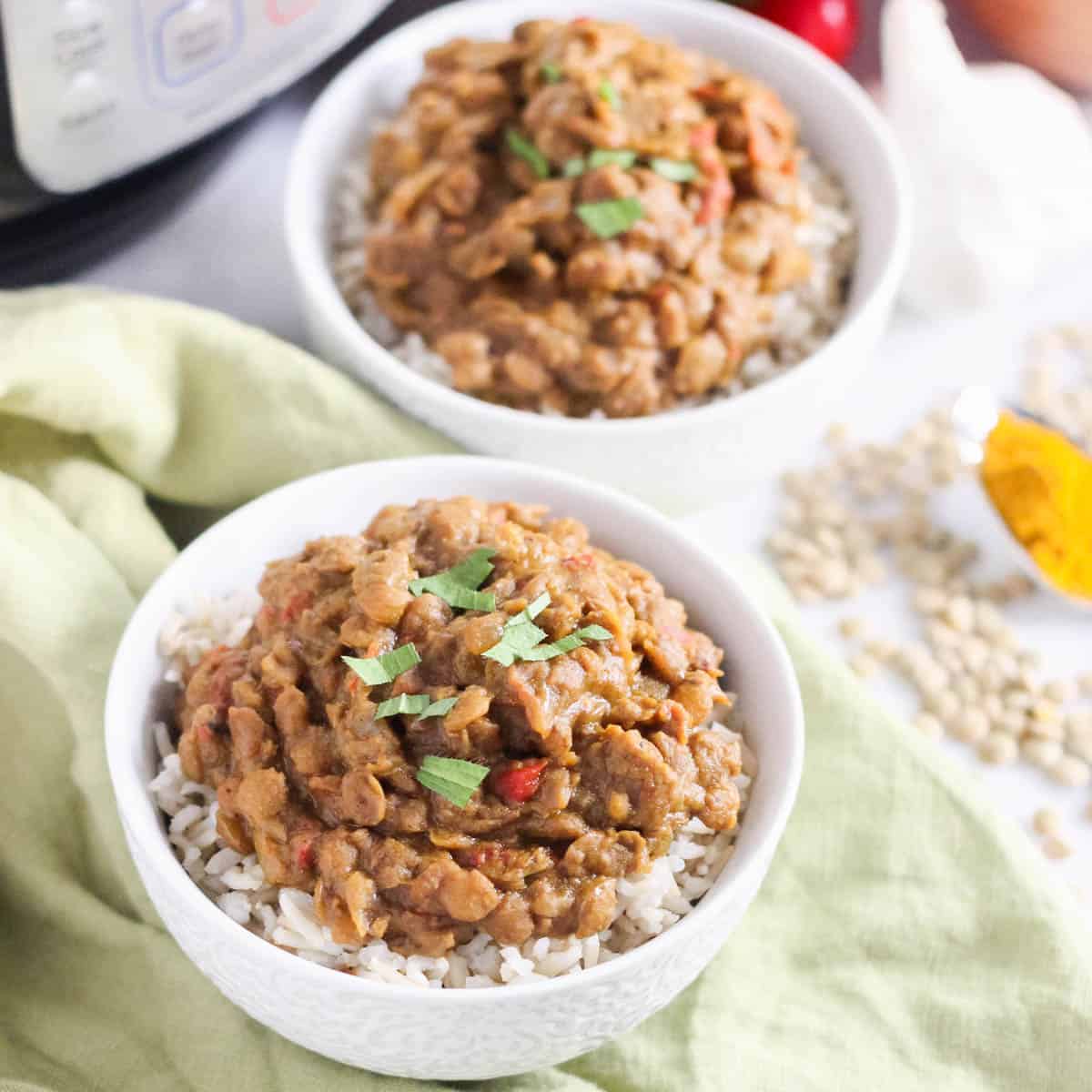 two small bowls of brown rice and lentil curry topped with parsley on a green napkin with dried lentils and pressure cooker in background.