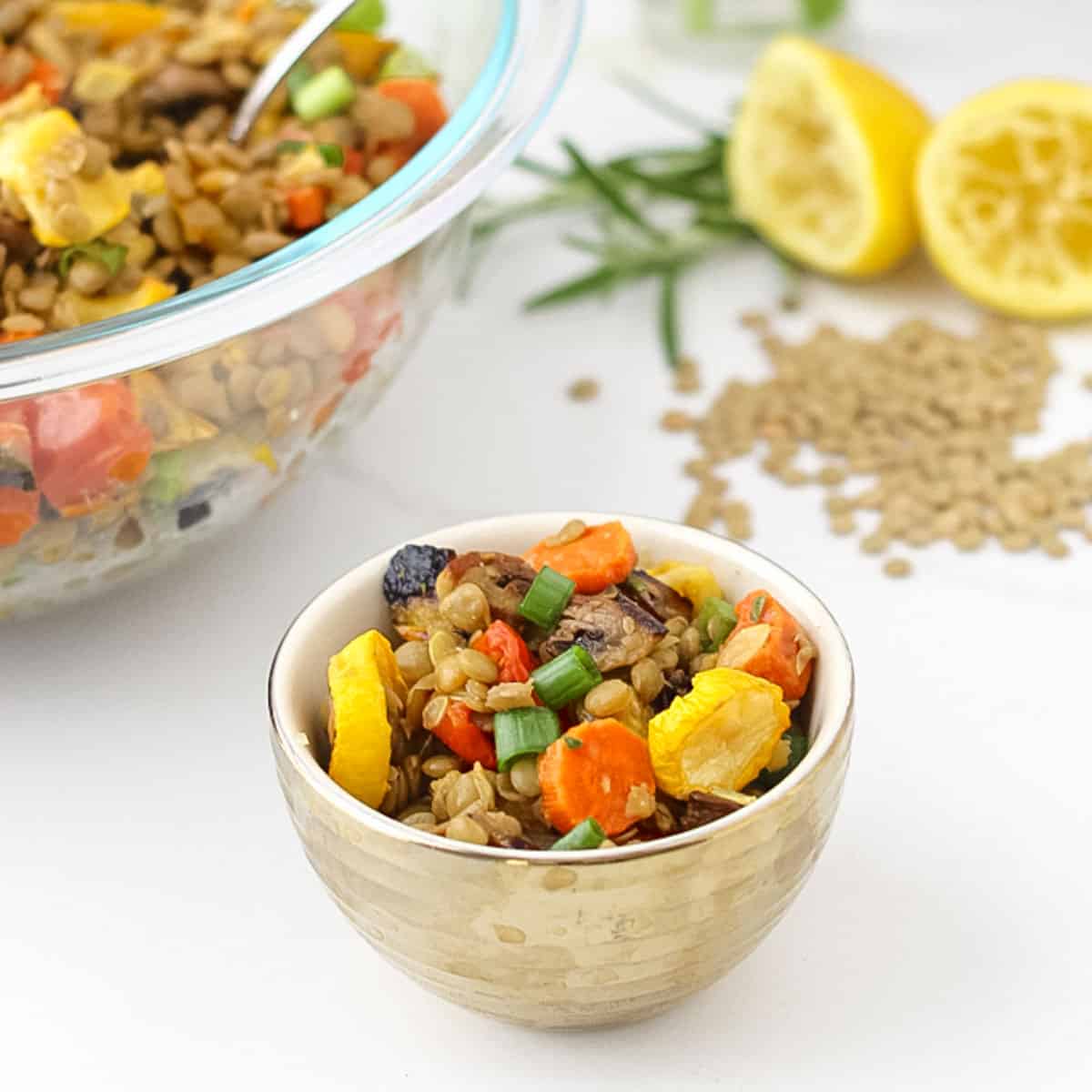 small gold bowl with lentil and roasted vegetable salad next to a big glass bowl full of salad, fresh rosemary, and lemons.