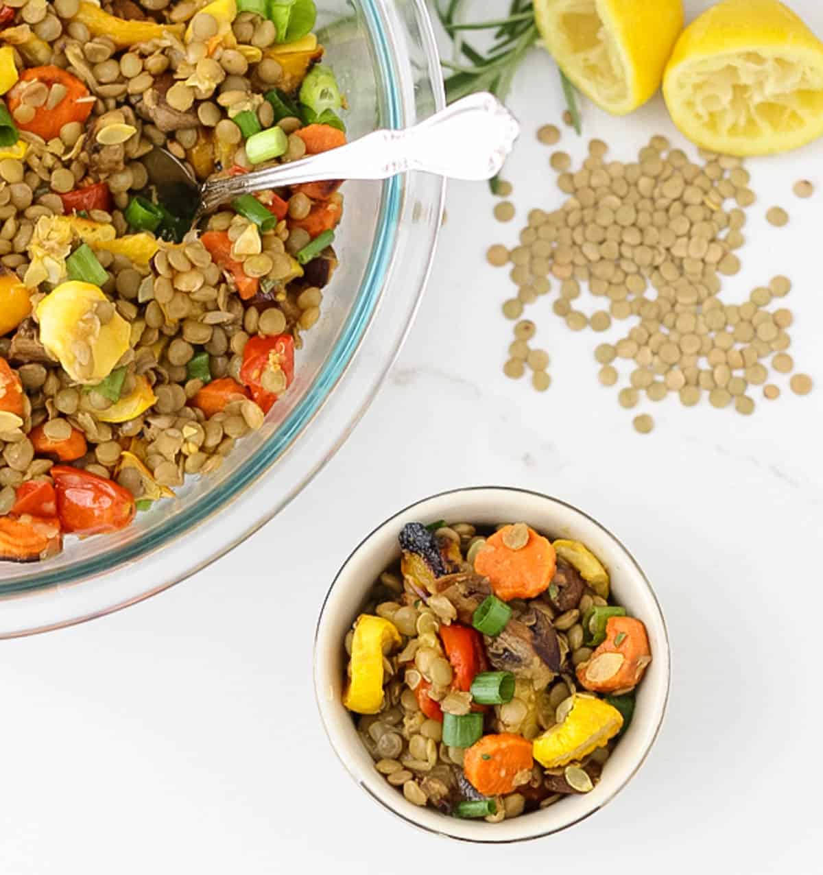 small bowl and large bowl of lentil salad with roasted veggies next to scattered green lentils, lemon, and fresh rosemary.