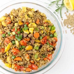 glass bowl of roasted vegetable lentil salad next to fresh rosemary, lemon, and scattered green lentils.