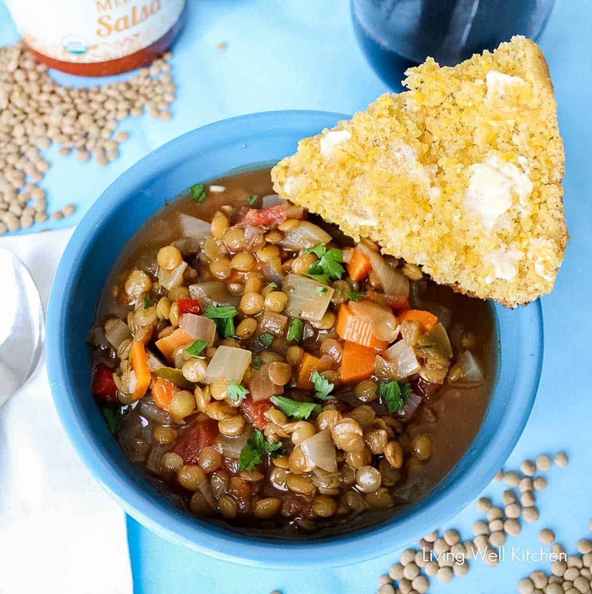 blue bowl of lentil soup with slice of buttered cornbread on a blue placemat surrounded by scattered green lentils