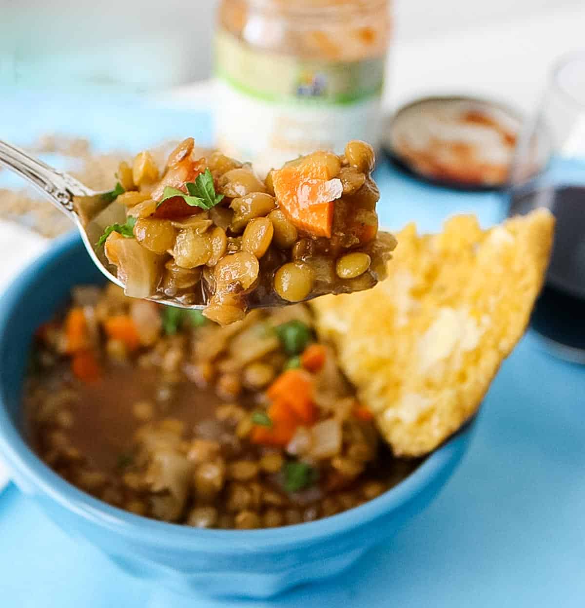 silver spoon with lentil soup over a blue bowl of soup on a blue placemat next to a glass of red wine and a jar of salsa.