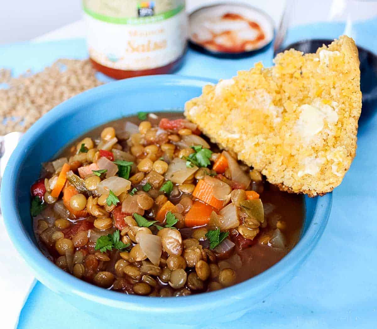 blue bowl full of lentil soup and a slice of buttered cornbread on a blue placemat with salsa, scattered green lentils, and a glass of red wine.