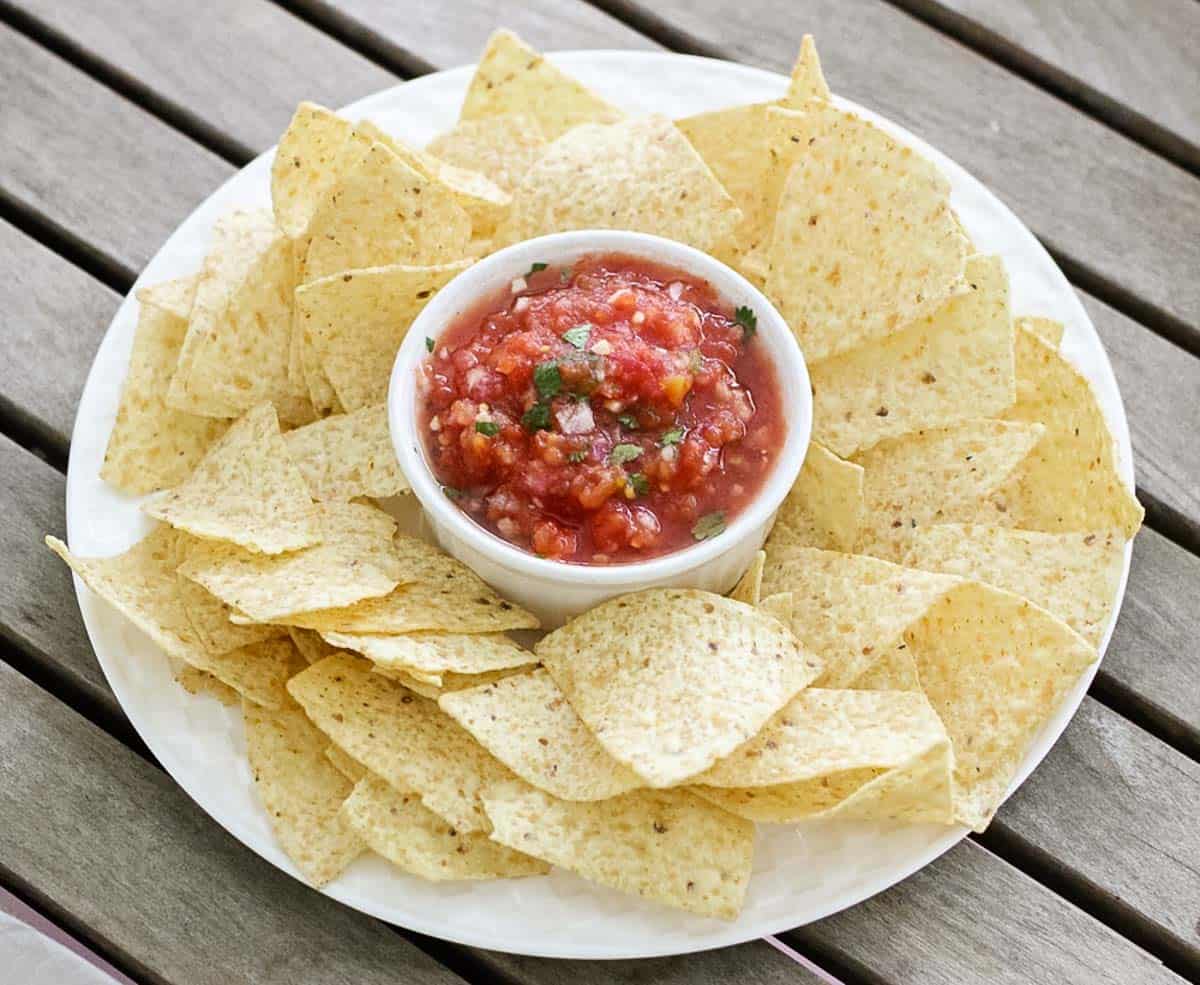 wooden table with white plate full of chips and a bowl of homemade low sodium salsa.