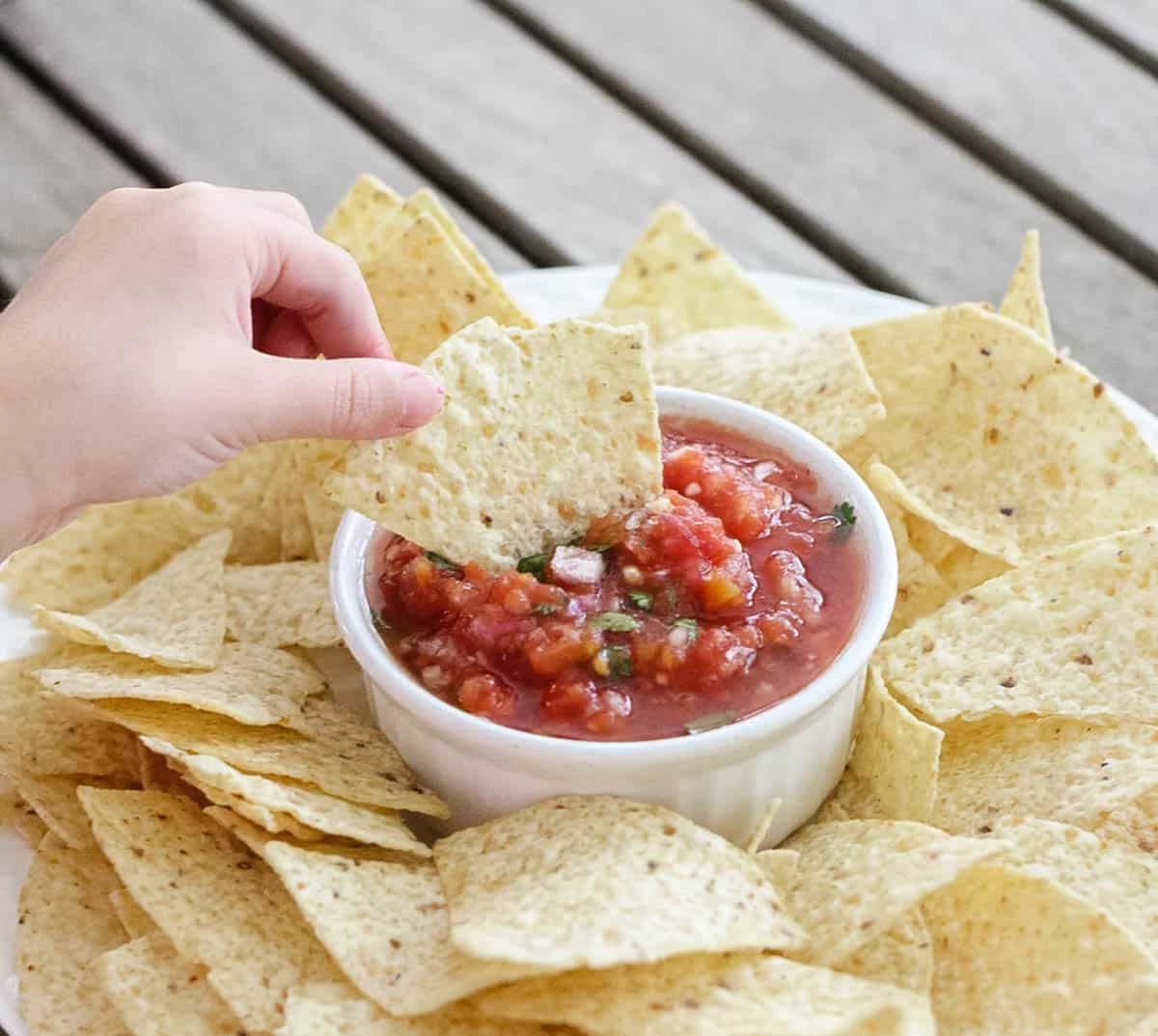 hand dipping a chip into homemade salsa recipe with canned diced tomatoes in a bowl with a plate of chips.