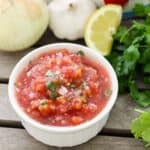 white bowl with tomato salsa next to fresh cilantro, garlic, onion, and lemon on a wooden table.