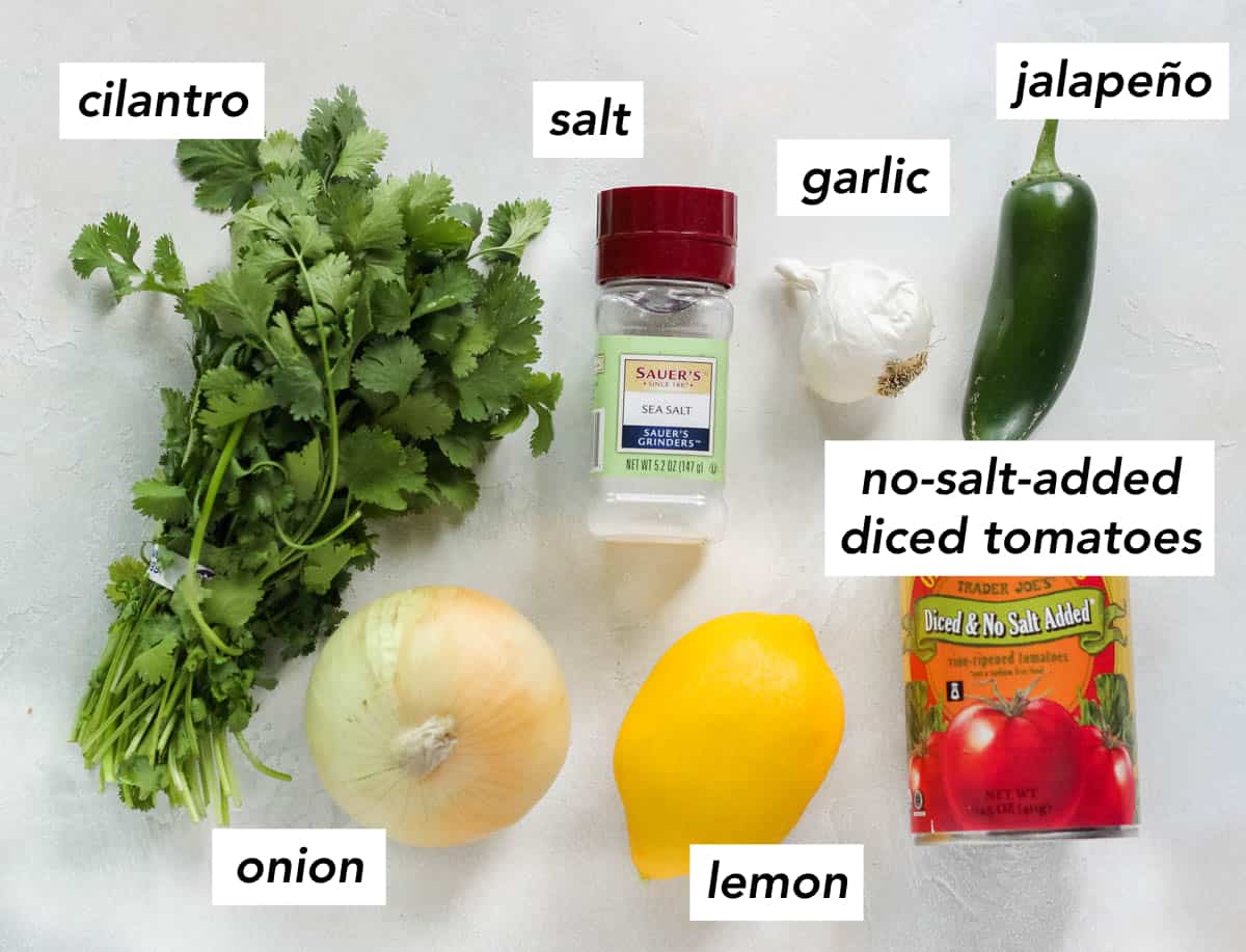 white counter with fresh cilantro, yellow onion, lemon, can of no salt added diced tomatoes, salt shaker, head of garlic, and fresh jalapeno with text overlay describing ingredients.