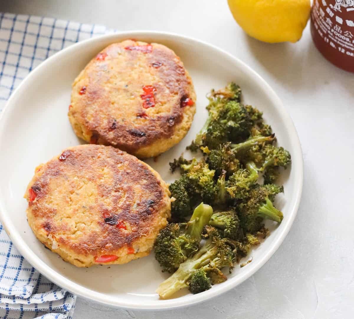 blue and white checkered napkin next to a plate with two salmon patties and roasted broccoli, with a lemon and sriracha sauce in background.