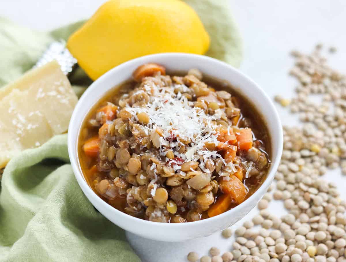 scattered green lentils, green napkin, parmesan cheese, and a white bowl of lentil soup topped with cheese.