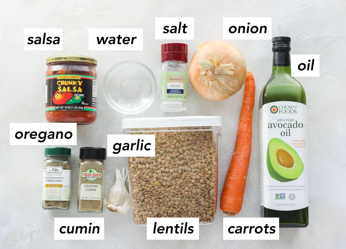 jar of salsa, bowl of water, salt, yellow onion, carrot, bottle of avocado oil, container of lentils, head of garlic, ground cumin, dried oregano on a white counter with text overlay labeling each ingredient.