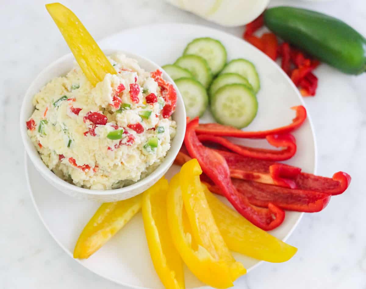 yellow bell pepper in a bowl of pimento cheese spread with sliced veggies on a plate.