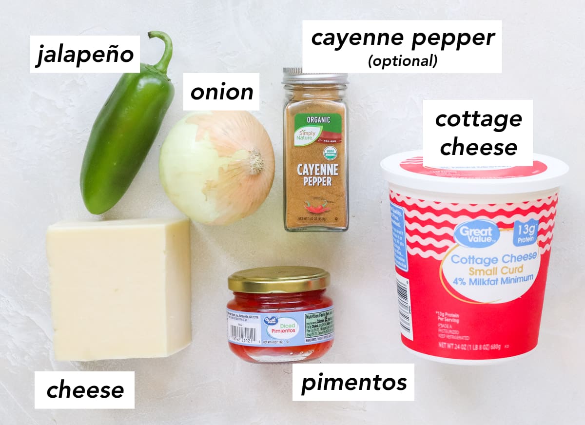 white counter with jalapeno, block of cheddar, jar of pimientos, container of cottage cheese, bottle of cayenne pepper, and a yellow onion.
