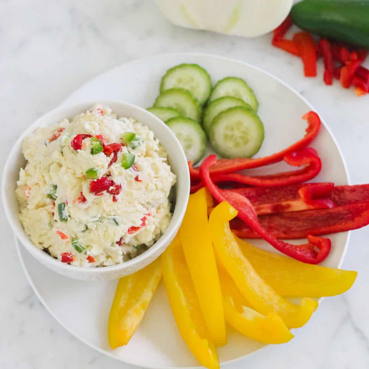 white plate with yellow and red bell pepper slices, cucumber slices, and a bowl of pimento cheese with jalapeno.