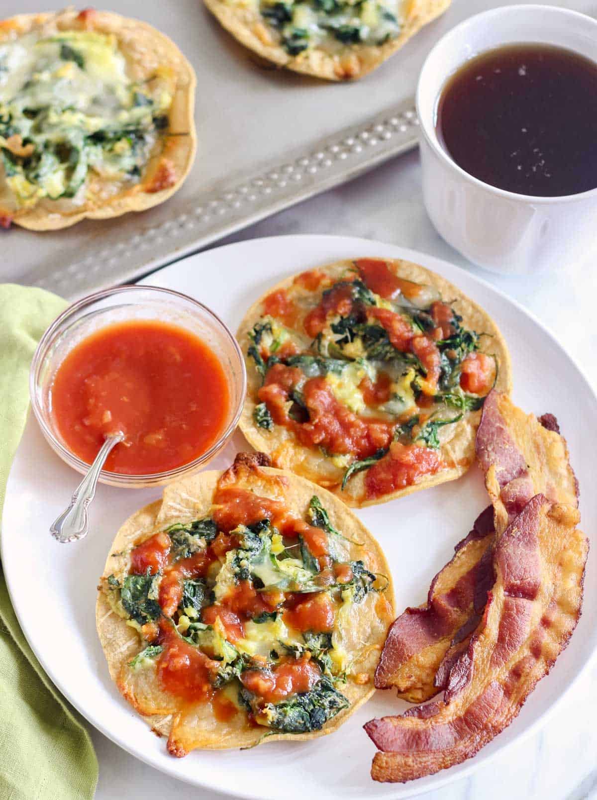white plate with two breakfast tostadas, slice of bacon, bowl of salsa next to a cup of tea, green napkin, and baking sheet with extra tostadas.