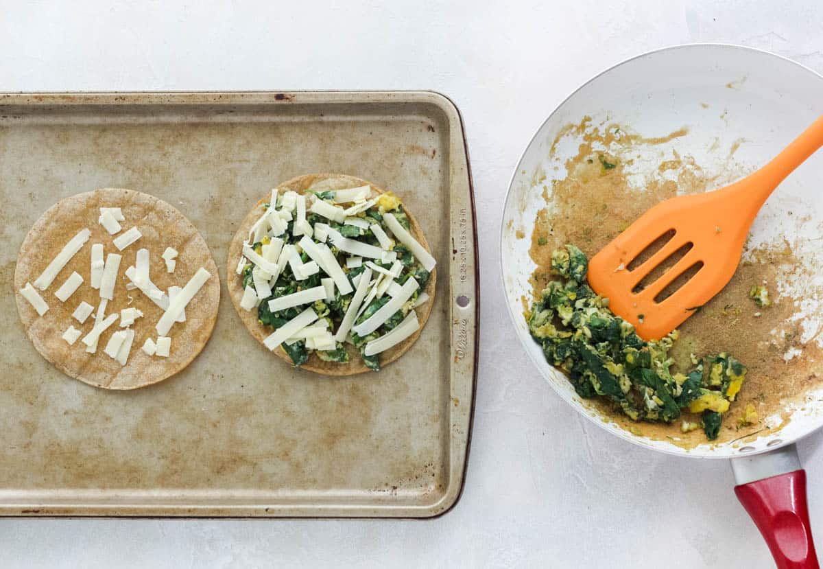 baking sheet with two homemade tostada shells one topped with cheese and the other topped with scrambled eggs and spinach, next to a skillet with spinach scrambled eggs and an orange spatula.