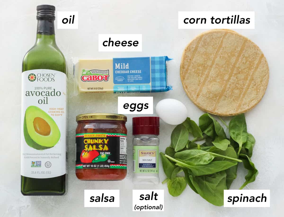 bottle of avocado oil, jar of salsa, salt shaker, handful of fresh spinach, egg, corn tortillas, block of cheddar cheese on white counter with text overlay describing ingredients.