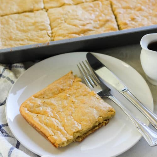 slice of baked protein pancakes on a white plate with a fork and knife next to a baking dish with sheet pan pancakes.