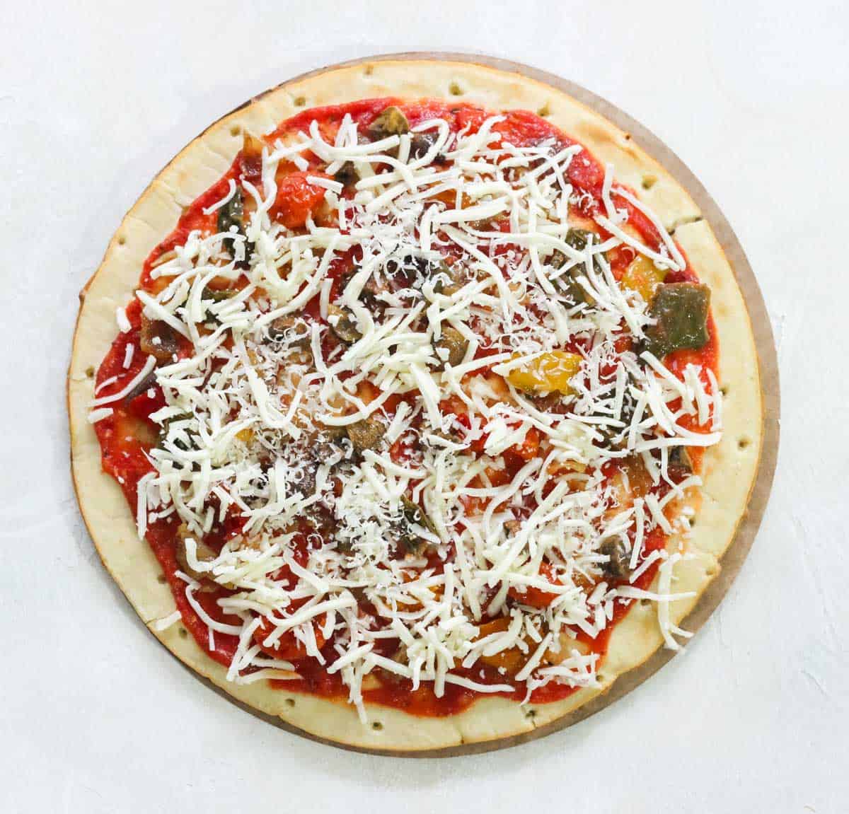 white counter with an unbaked pizza crust topped with pizza sauce, roasted veggies, and cheese before baking.