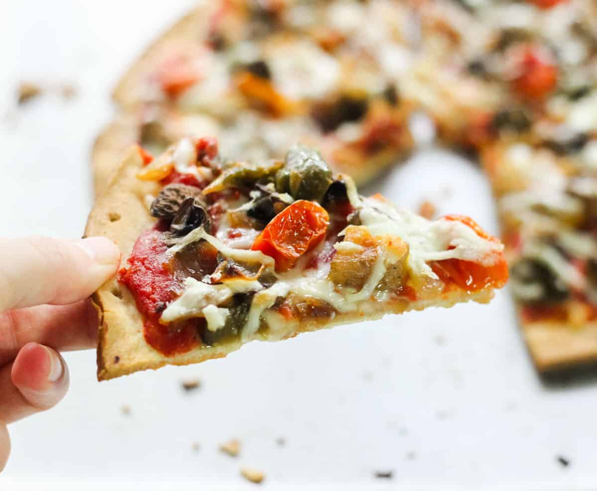 hand holding a slice of roasted vegetable pizza with a pizza in the background.