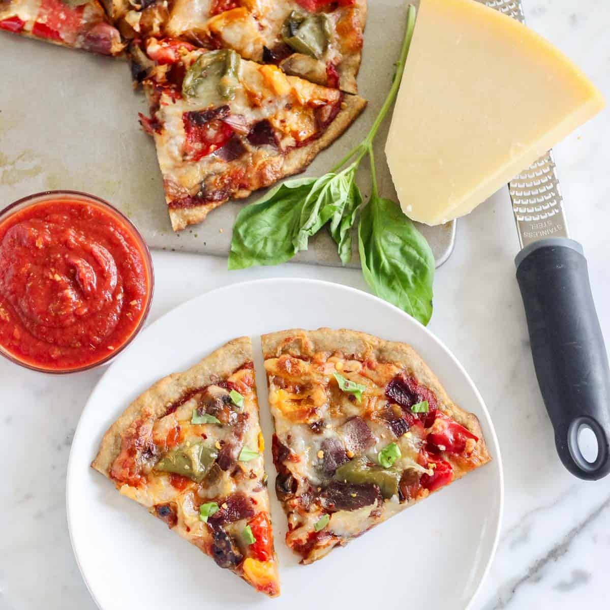 white plate with two slices of roasted vegetables pizza next to parmesan, fresh basil, a microplane, a bowl of pizza sauce, and a baking sheet with pizza.