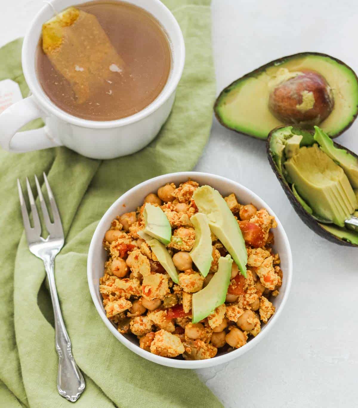 white bowl with scrambled tofu topped with avocado next to a fork, green napkin, cup of tea, and a cut avocado.