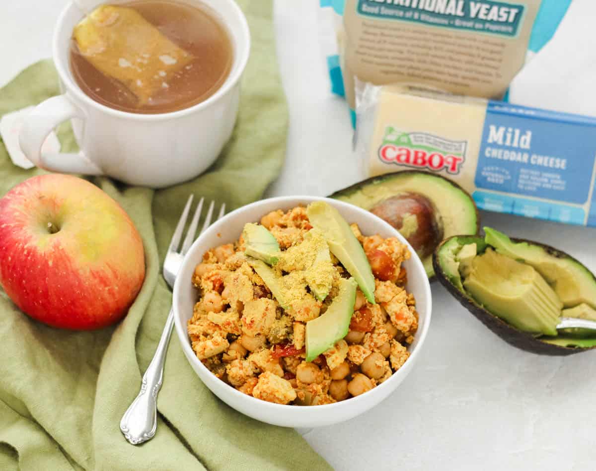 tofu scramble in a bowl topped with avocado slices and nutritional yeast, next to a cup of tea, an apple, a fork, avocado, block of cheese, and bag of nutritional yeast.