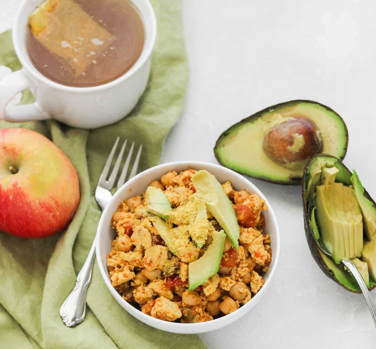 southwest tofu scramble in a bowl topped with avocado slices and nutritional yeast next to a cup of tea, an apple, a fork, and an avocado.