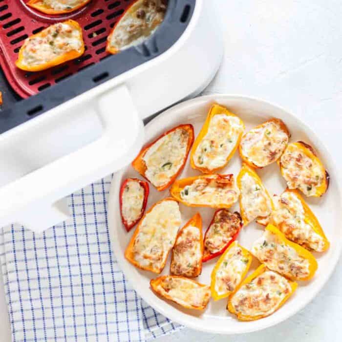 plate with stuffed mini bell peppers next to an air fryer with stuffed peppers inside and a blue and white napkin.