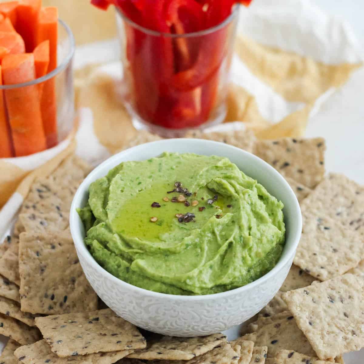 white bowl of spinach hummus surrounded by crackers and cups of sliced veggies.