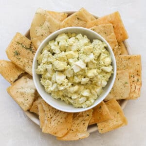 bowl of egg salad hummus surrounded by pita chips on a white counter.