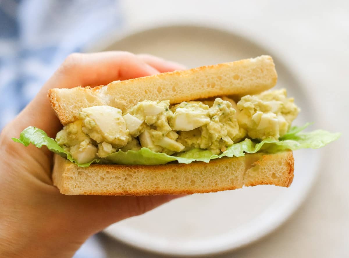hand holding egg hummus sandwich with a plate in the background.