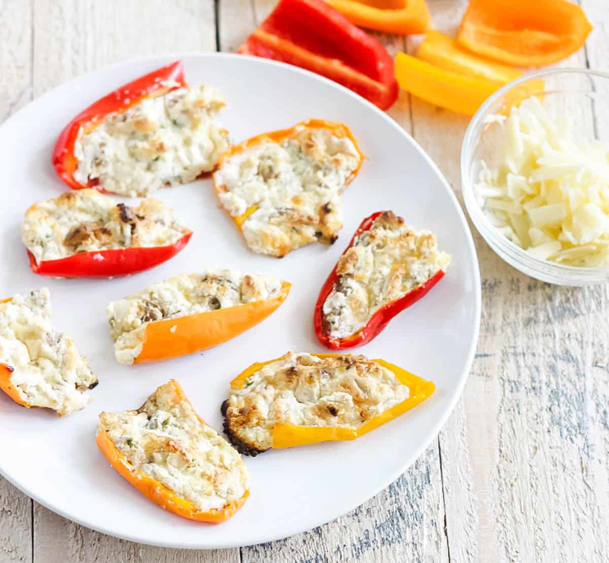 white plate of cream cheese stuffed mini peppers next to a bowl of grated cheese and halved baby bell peppers on a wooden table.