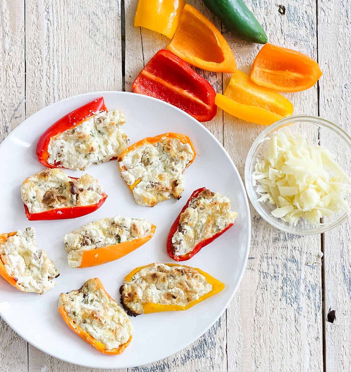 white plate with mini peppers stuffed with cream cheese surrounded by jalapeños, baby bell peppers, and a bowl of grated cheese.