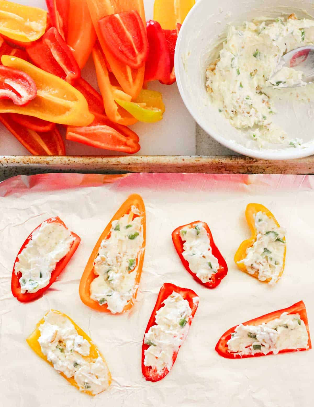 halved mini bell peppers, a bowl of cream cheese, and a foil lined baking sheet with cream cheese stuffed bell peppers.