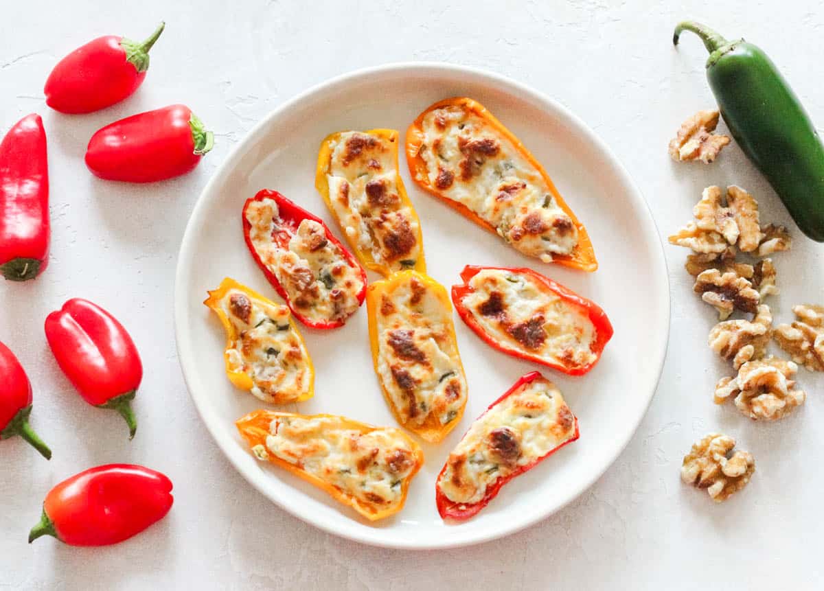 red mini bell peppers, a white plate with baked cream cheese stuffed bell peppers, a jalapeno, and walnuts.