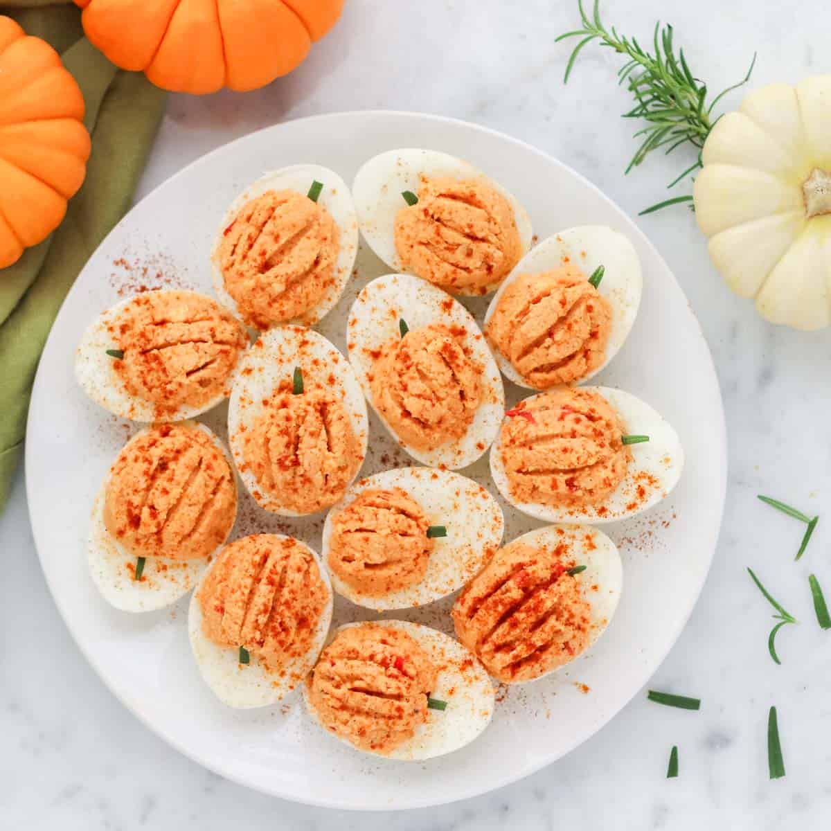 white plate with pumpkin deviled eggs on a marble kitchen counter surrounded by chopped rosemary plus small orange and white pumpkins.