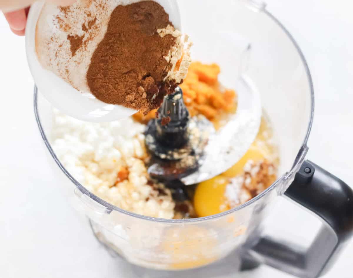 bowl of spices being poured into a bowl of a food processor with cottage cheese, eggs, and oats.