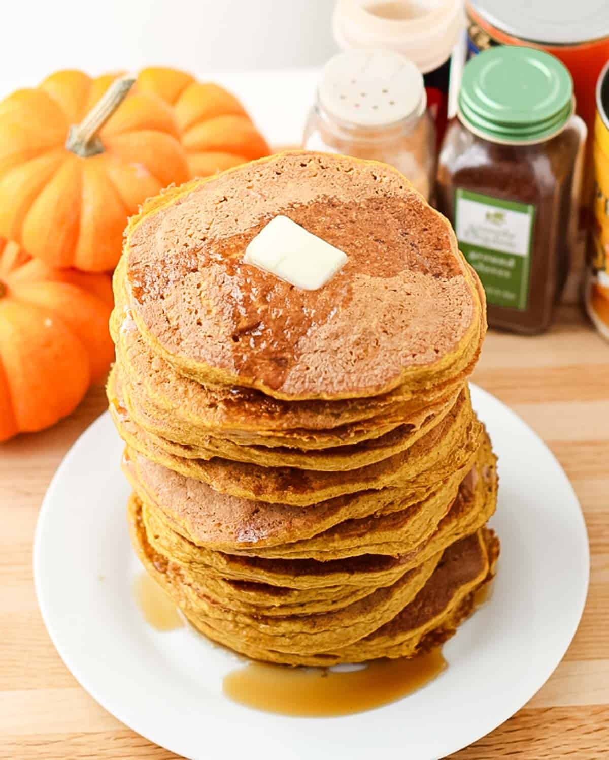 plate of pumpkin pancakes stacked high and topped with butter and maple syrup surrounded by spices and fresh pumpkins.